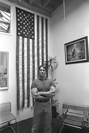 Arnold Schwarzenegger standing in his office with USA flag and photos on the wall.